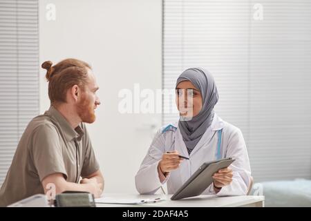 Weibliche Arzt in Hijab mit digitalen Tablette während der Diskussion Methoden Der Behandlung mit ihrer Patientin am Tisch im Krankenhaus Stockfoto