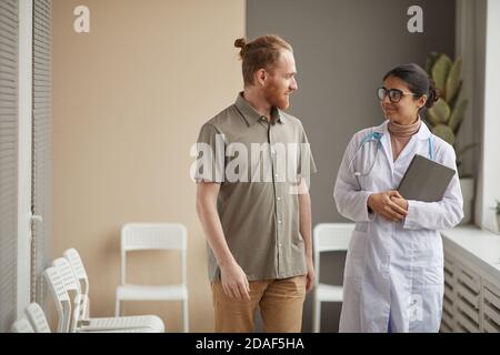 Junge Ärztin im weißen Mantel im Gespräch mit männlichen Patienten Während sie im Krankenhaus den Korridor entlang gehen Stockfoto