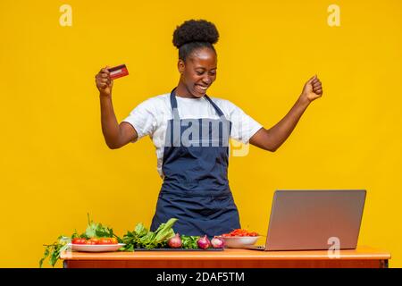 Aufgeregt afrikanischen Koch freut sich in der Mitte der Vorbereitung ein Essen, während sie ihre Kreditkarte und mit ihrem Laptop Computer Stockfoto
