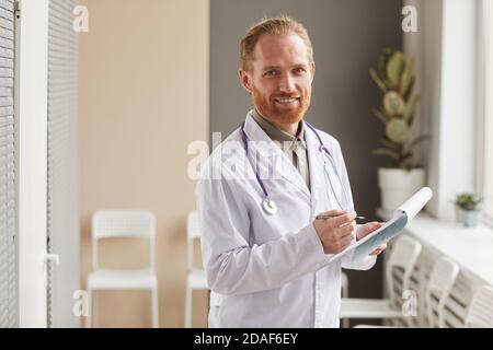 Porträt von bärtigen männlichen Arzt in weißem Mantel hält medizinische Karte und lächelnd auf die Kamera, die am Flur steht Krankenhaus Stockfoto