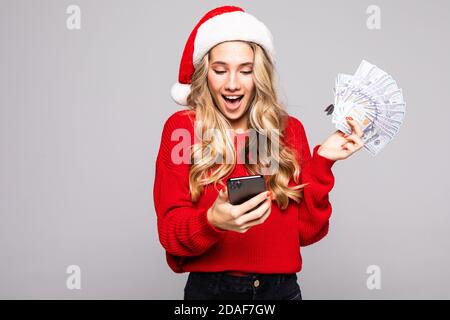 Porträt einer jungen überraschten Frau in rotem weihnachtshut Halten Bündel von Geldscheinen während der Verwendung von Handy über Weißer Hintergrund Stockfoto