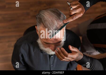 Afroamerikanischer Barbier mit tätowierten Händen kämmt das Kopfhaar eines grauhaarigen, bärtigen kaukasischen Mannes, von oben gesehen. Stockfoto