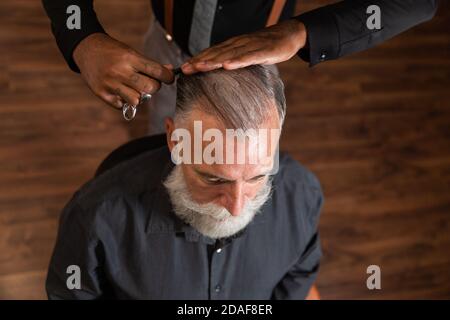 Afroamerikanischer Barbier mit tätowierten Händen kämmt das Kopfhaar eines grauhaarigen, bärtigen kaukasischen Mannes, von oben gesehen. Stockfoto