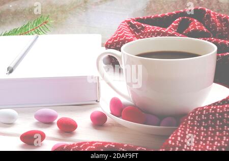 Eine weiße Tasse Kaffee mit bunten Süßigkeiten steht auf Eine hölzerne Fensterbank neben einem warmen Schal und White Book Stockfoto