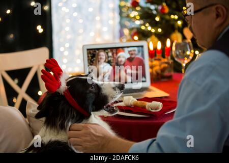 Reifer Mann feiert Weihnachten mit seinem Hund sitzt am servierten Feiertagstisch mit Laptop. Personen grüßen ihre Freunde bei einem Videoanruf mit einer Webcam. Stockfoto