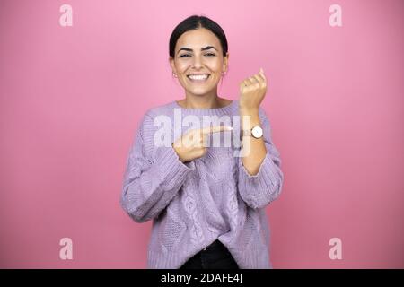 Schöne Frau trägt einen lässigen violetten Pullover auf rosa Hintergrund Lächelnd und mit dem Zeiger auf seine Uhr, weil es spät ist Stockfoto