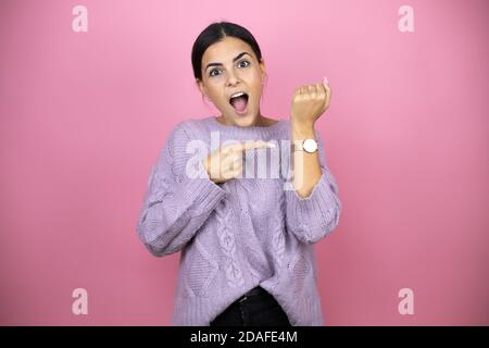 Schöne Frau trägt einen lässigen violetten Pullover auf rosa Hintergrund Überrascht und zeigt seine Uhr, weil es spät ist Stockfoto