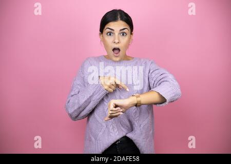 Schöne Frau trägt einen lässigen violetten Pullover auf rosa Hintergrund Überrascht und zeigt seine Uhr, weil es spät ist Stockfoto