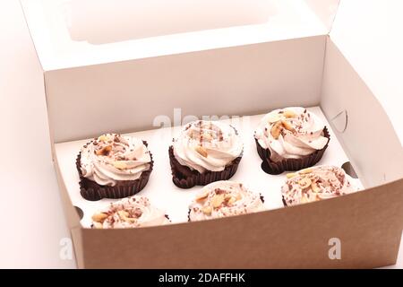 Sechs Schokoladen-Muffins mit weißer Sahne und Erdnüssen in einer offenen Schachtel. Süßigkeiten auf einem weißen Tisch Stockfoto