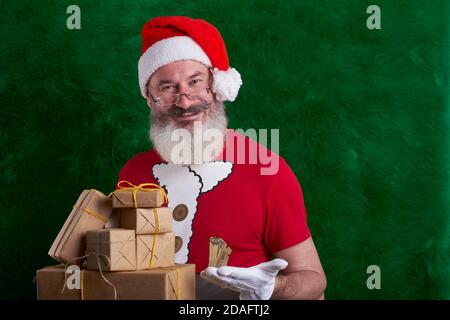 Reifer bärtiger Mann trägt Weihnachtsmütze mit vielen Geschenken in der Hand, Santa lächelt und schaut auf Kamera, kopieren Raum Stockfoto