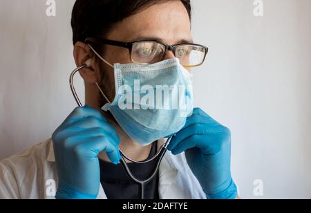 Ein junger, hübscher Arzt mit Brille und medizinischer Gesichtsmaske legt ein Stethoskop an, während er sich auf eine Patientenuntersuchung vorbereitet. Stockfoto