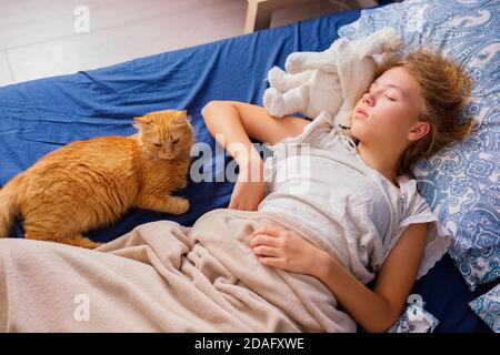 Ein Mädchen mit blonden Haaren schläft morgens im Bett neben einer flauschigen roten Katze. Weicher, freier Fokus. Stockfoto