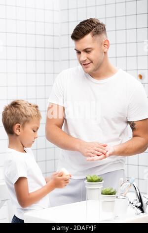 Positiver Vater, der sich die Hände wascht und den Sohn mit Seife ansieht In der Nähe des Waschbeckens im Badezimmer Stockfoto