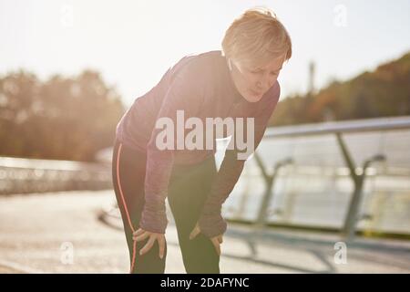 Übertreiben Sie es nicht. Aktive reife Frau in Sportbekleidung, müde und erschöpft, eine Pause nach dem Laufen im Freien an einem sonnigen Tag Stockfoto