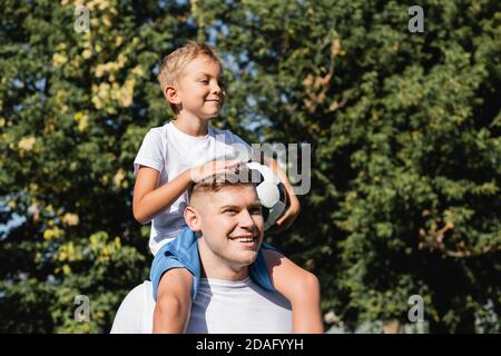 Lächelnder Sohn mit Ball reiten Huckepack auf glücklichen Vater in parken Sie auf unscharfem Hintergrund Stockfoto