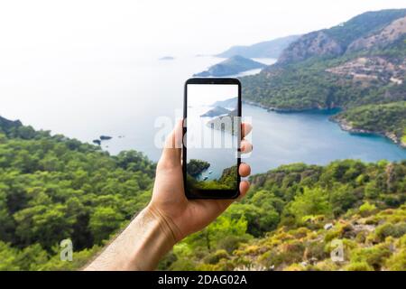 Eine Hand mit dem Smartphone, die auf einer Reise Bilder von der Naturlandschaft, dem Meer und den Bergen macht. Telefon-, Internet- und Mobilfunkkonzept. Beeindrucken Stockfoto