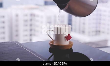 Gießen von heißem Wasser aus einem Wasserkocher zu Keramik-Becher mit einem Teebeutel, selektive Fokus und Nahaufnahme Blick auf das Büro. Stockfoto