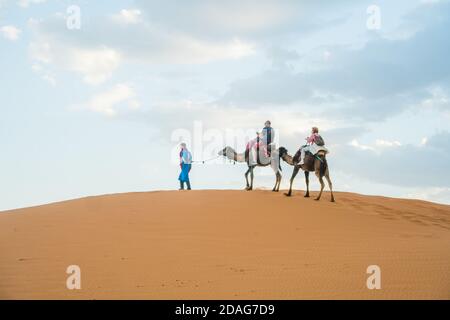 Merzouga, Marokko - APRIL 29 2019: Marokkaner wandern und führen zwei Kamele mit alten Touristen in der Sahara Wüste bei Sonnenuntergang Stockfoto