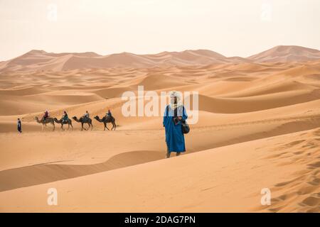 Merzouga, Marokko - APRIL 29 2019: Marokkaner stehen im Sand in der Sahara und blicken auf den Horizont mit Kamelen im Hintergrund Stockfoto