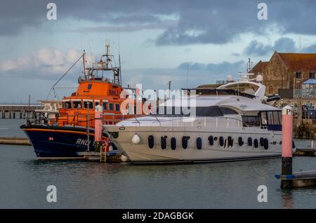 Das RNLI-Rettungsboot in yarmouth auf der Insel wight neben einem Luxus-Motorboot oder einer Motoryacht an einem Herbstabend im stimmungsvollen Licht. Stockfoto