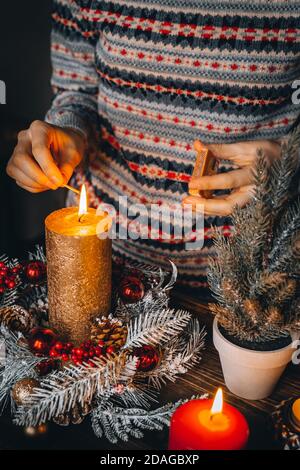 Frau Hände Blitz große goldene Kerze mit Streichhölzern in Weihnachtskranz. Weihnachten Innendekoration auf rustikalem Holzhintergrund. Hygge, Ornamente, Kerze Stockfoto