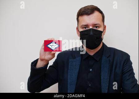 Mann tragen schwarz formell und schützen Gesichtsmaske, halten Arkansas Flagge Karte isoliert auf weißem Hintergrund. USA Coronavirus Covid Länderkonzept. Stockfoto