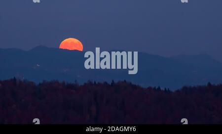 Riesiger oranger Vollmond, der in den Nachthimmel dahinter aufsteigt Ein Bergrücken mit Hügelschichten im Vordergrund Eine mystische Atmosphäre schaffen Stockfoto