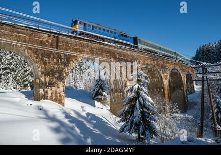 Steinviadukt (Bogenbrücke) auf Eisenbahn durch schneebedeckten Tannenwald und Lokomotive mit Personenzug. Schnee driftet am Wegesrand und Reif Stockfoto