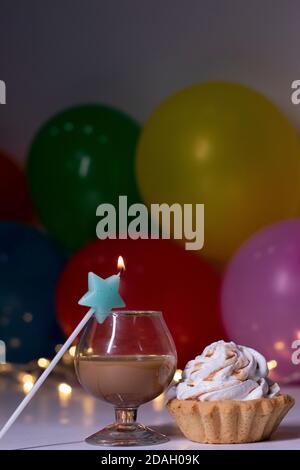 cupcake mit einer blauen Sternkerze und einem Glas mit einem braunen Flüssiglikör. Auf dem Hintergrund von bunten Ballons und Lichter. Party, Geburtstagskonzept. Speicherplatz kopieren. . Hochwertige Fotos Stockfoto