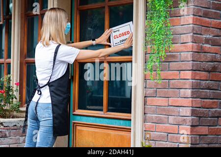 Kaukasische Kellnerin Frau trägt medizinische Maske tut UNS LEID, WIR SIND GESCHLOSSEN Pandemie-Zeichen auf Café-Fenster. Coronavirus-Pandemie. Abschaltung der Regierung von Stockfoto