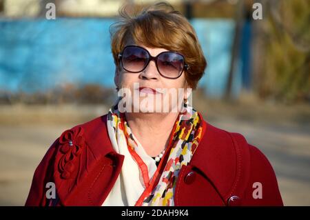 Porträt von stilvollen Senior Frau in einem hellen roten Mantel und Sonnenbrille freundlich und aufrichtig lächelnd, Blick auf die Kamera Stockfoto