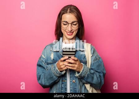 Lächelndes junges Mädchen in Denim Jacke schaut auf das Telefon auf einem rosa Hintergrund. Stockfoto