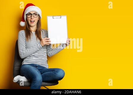 Die junge Frau in einem weihnachtsmann-Hut sitzt auf einem Stuhl und hält eine Zwischenablage mit einem leeren Blatt Papier auf einem gelben Hintergrund. Banner Stockfoto