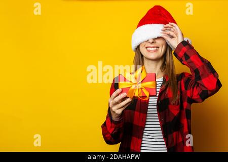 Junge Frau gucken aus unter einem weihnachtsmann Hut und Ein rot kariertes Hemd und hält ein Geschenk in ihr Hände auf gelbem Hintergrund Stockfoto