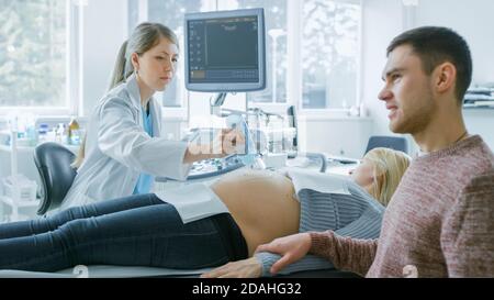 Im Krankenhaus legt Geburtshelferin Gel auf den Bauch einer schwangeren Mutter, bereit, Ultraschall-Screening zu tun. Ehemann Unterstützt Fleißig Seine Stockfoto