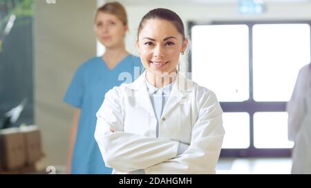 Schöne junge Ärztin geht durch den Flur des Krankenhauses, sie hält an, kreuzt Arme und charmant Smiles. Professionelle Mitarbeiter bei der Arbeit. Stockfoto