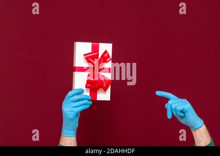 Neujahr 2021. Weihnachtsfeier, Schutz vor Coronavirus. Mann Hände tragen medizinische Gummi blaue Handschuhe zeigen die Geschenk-Box.Red st Stockfoto