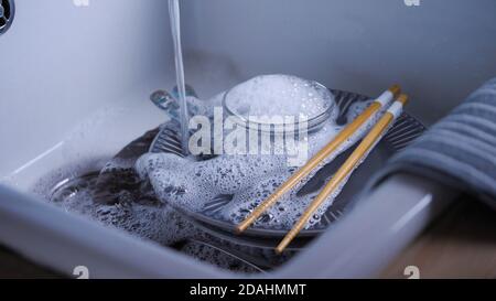 Hausreinigung Routine. Wasser strömt auf das Geschirr in der Küchenspüle. Geschirr von Hand waschen. Blaue Gerichte und chinesische Essstäbchen. Haus Wasser Energie sparen. Stockfoto