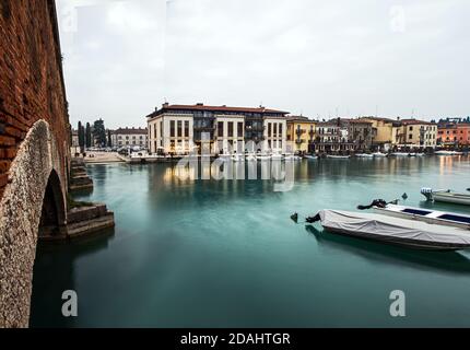 Landschaftlich schöne Aussicht auf Plattform im Seekanal mit Schwan, Boot und Häuser und Hotel im Hintergrund während der blauen Stunde, Peschiera del Garda, Verona, Italien Stockfoto