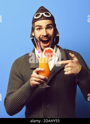 Schöner bärtiger Flieger Mann mit langem Bart auf lächelndem Gesicht hält Glas alkoholischen Cocktail in grau gestrickten Pullover mit Hut und Brille, auf blauem Studio-Hintergrund Stockfoto