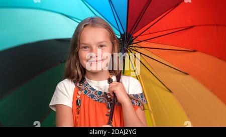 Lächelndes Mädchen in Mutter Sundress mit Perlen hält offen hellen bunten Regenschirm. Kaukasisches Kind schaut auf die Kamera und lächelt Stockfoto