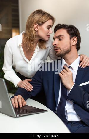 Leidenschaftliches Paar flirtet im Büro. Geschäftsmann sieht auf Frau, die ihn von hinten berührt, während sie in der Nähe des Arbeitsplatzes steht. Verführerische Konzept Stockfoto