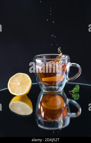 Ein Glas Tasse mit schwarzem Tee, ein Spritzer in der Tasse von fallenden Zitrone, Reflexion auf schwarzem Hintergrund, Zitrone und Minze Blatt auf dem Tisch angeordnet Stockfoto