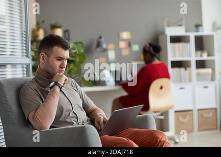 Seriöser junger Mann, der sich auf seine Online-Arbeit konzentriert Sessel und Arbeit am Laptop mit Frau mit Laptop in Der Hintergrund Stockfoto