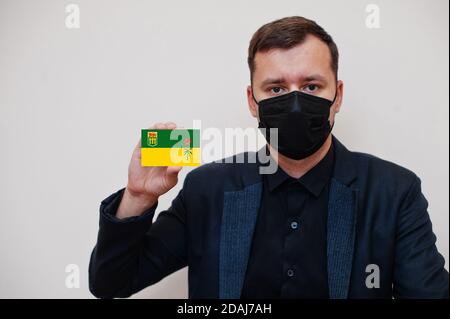 Mann tragen schwarz formell und schützen Gesichtsmaske, halten Saskatchewan Flagge Karte isoliert auf weißem Hintergrund. Kanada Provinzen Coronavirus Covid Land Konz Stockfoto