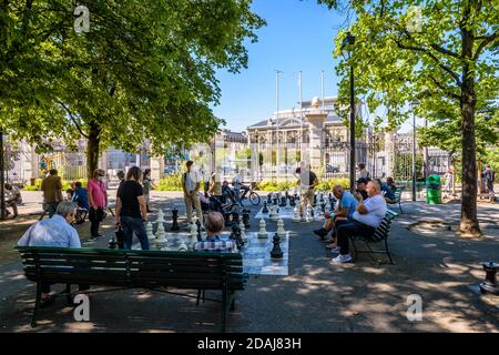 Auf riesigen Schachbrettern im Schatten im Park der Bastionen in Genf spielen die Leute Schach, und die Zuschauer sitzen auf Bänken herum. Stockfoto