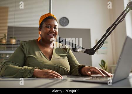 Porträt einer glücklichen afrikanischen Frau, die während des Sitzens die Kamera anlächelt Am Tisch mit Laptop und führender Live-Übertragung Stockfoto