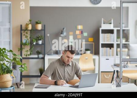 Junger Geschäftsmann sitzt am Tisch vor dem Laptop und schriftlich Pläne in seinem Notizblock er arbeitet Im Büro Stockfoto