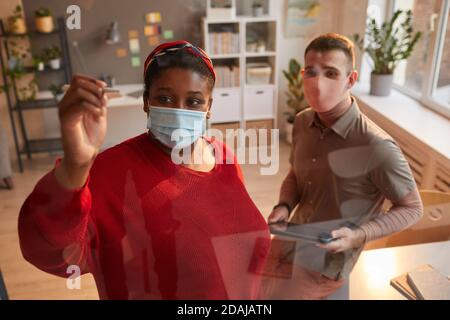 Afrikanische Frau in Maske schreiben auf der Glaswand sie Arbeiten über Projekt zusammen mit Mann im Büro Stockfoto
