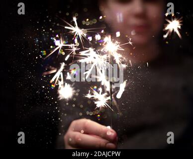 Weihnachtsfeier. Ein Kind hält ein brennendes Feuerwerk auf einem glänzenden Hintergrund mit leuchtend goldenen Weihnachtslichtern. Dunkles Konzept. Stockfoto